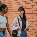 Cheerful multiracial female students wearing casual clothes carrying backpack and notebooks near college brick wall while discussing plans and looking at each other with smiles