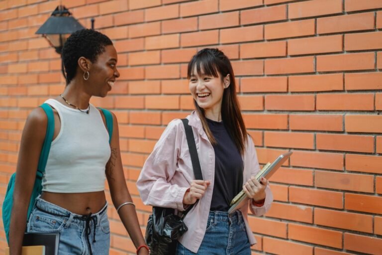 Cheerful multiracial female students wearing casual clothes carrying backpack and notebooks near college brick wall while discussing plans and looking at each other with smiles